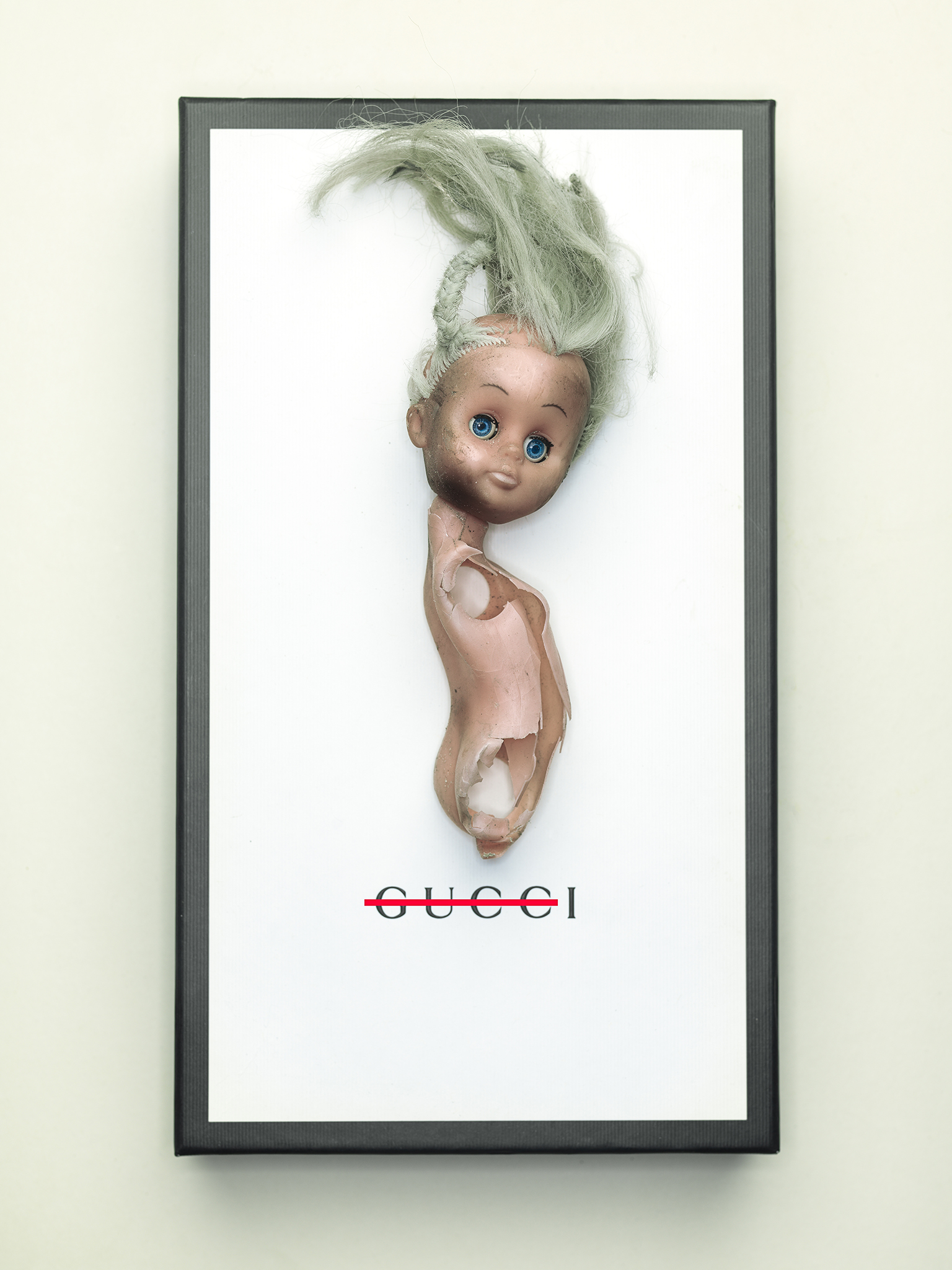 broken doll with blue eyes on a Gucci box conceptual fine art by Thabit